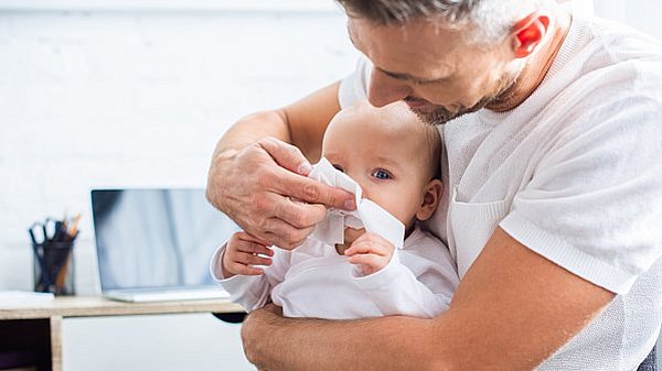 Kinder sind besonders anfällig für Erkältungen und Schnupfen. Ein Vater hält seinen Säugling auf dem Arm und putzt ihm die Schnupfennase mit einem Papiertaschentuch. © LightFieldStudios / iStock