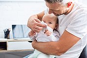 Ein Vater hält seinen Säugling auf dem Arm und putzt ihm die Schnupfennase mit einem Papiertaschentuch.