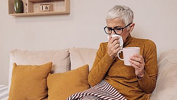 Eine ältere Frau mit Schnupfen sitzt auf dem Sofa mit einer Decke  über den Beinen und einem Glas Tee in der Hand. Mit der anderen Hand schnäuzt sie sich die Nase.