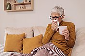 Eine ältere Frau mit Schnupfen sitzt auf dem Sofa mit einer Decke  über den Beinen und einem Glas Tee in der Hand. Mit der anderen Hand schnäuzt sie sich die Nase.