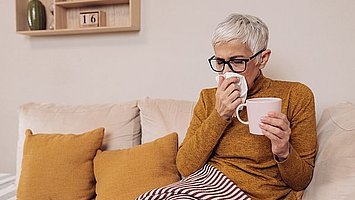 Eine ältere Frau mit Schnupfen sitzt auf dem Sofa mit einer Decke  über den Beinen und einem Glas Tee in der Hand. Mit der anderen Hand schnäuzt sie sich die Nase.