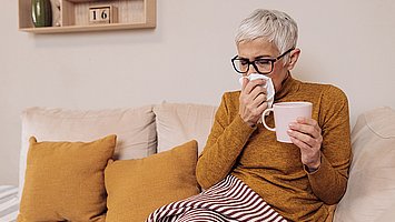 Eine ältere Frau mit Schnupfen sitzt auf dem Sofa mit einer Decke  über den Beinen und einem Glas Tee in der Hand. Mit der anderen Hand schnäuzt sie sich die Nase.