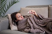Erkältete Frau unter Decke Fieber und oder erhöhte Temperatur: Eine Frau kontrolliert ihre Körpertemperatur mit einem digitalen Thermometer, um die Schwere ihrer grippalen Erkrankung zu beurteilen.